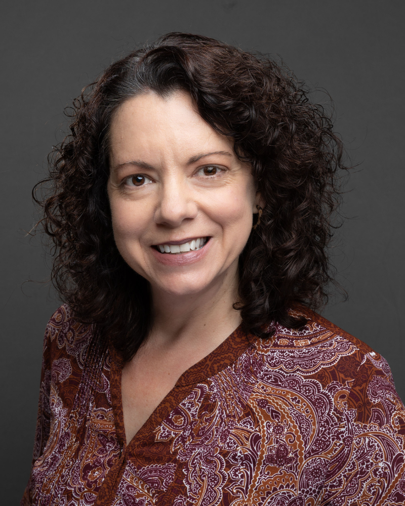 Headshot of a woman with curly dark hair smiling, wearing a patterned blouse against a gray background
